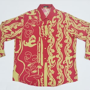 May include: A red button-down shirt with long sleeves and a collared neckline. The shirt features a repeating pattern of gold-colored abstract shapes and lines. The shirt has gold buttons and cuffs. The shirt is laid flat on a white surface.