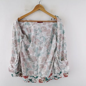 May include: A white, long-sleeved top with a mermaid pattern in pink and teal. The top has a wrap-around design with pockets and is hanging on a wooden hanger. The bottom hem features a contrasting border with mermaid illustrations.