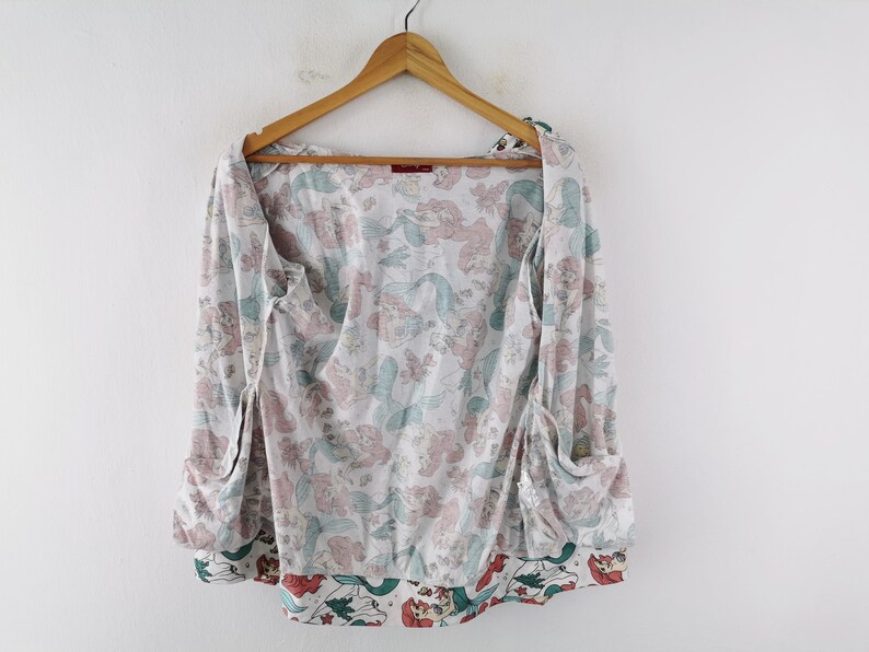 May include: A white, long-sleeved top with a mermaid pattern in pink and teal. The top has a wrap-around design with pockets and is hanging on a wooden hanger. The bottom hem features a contrasting border with mermaid illustrations.