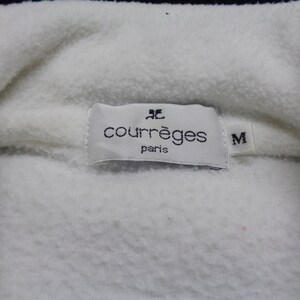 May include: Close-up of a white fleece garment with a label that reads "courr&egrave;ges paris" and the size "M". The fabric is soft and textured.