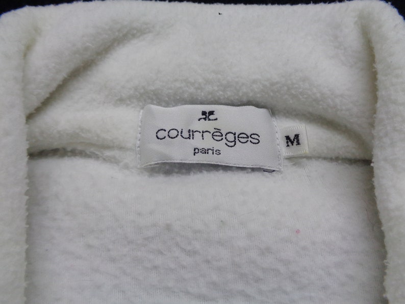 May include: Close-up of a white fleece garment with a label that reads "courr&egrave;ges paris" and the size "M". The fabric is soft and textured.