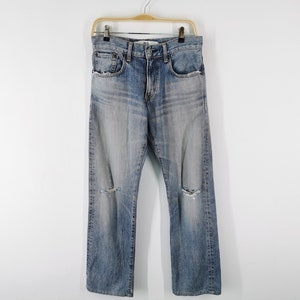 May include: Light blue denim jeans with distressed details, including knee rips. The jeans are hanging on a wooden hanger against a white background. The jeans have a classic five-pocket design.