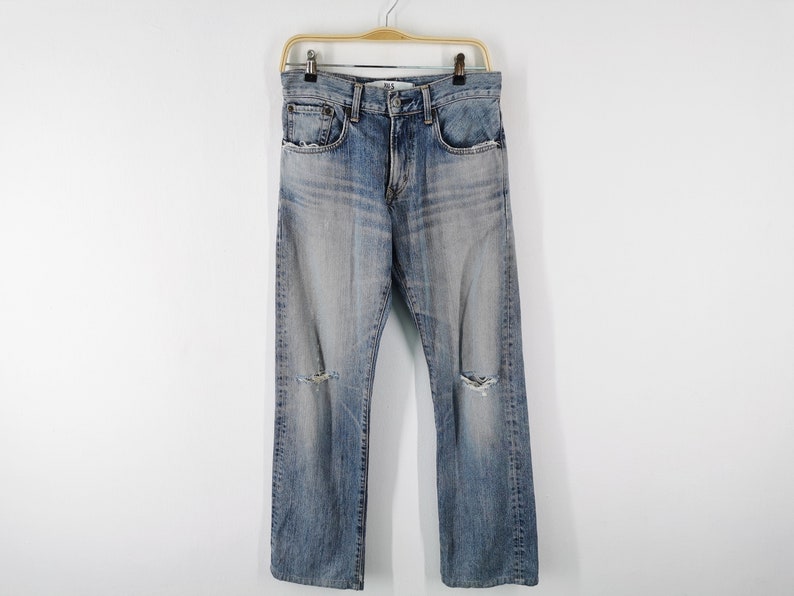 May include: Light blue denim jeans with distressed details, including knee rips. The jeans are hanging on a wooden hanger against a white background. The jeans have a classic five-pocket design.