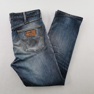 May include: Blue denim jeans with a classic five-pocket design. The jeans feature a faded wash and a leather patch on the back pocket with the "Wrangler" logo. The jeans are folded and laid flat, showcasing the details of the stitching and the overall fit.