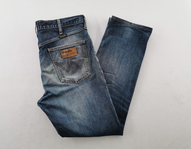 May include: Blue denim jeans with a classic five-pocket design. The jeans feature a faded wash and a leather patch on the back pocket with the "Wrangler" logo. The jeans are folded and laid flat, showcasing the details of the stitching and the overall fit.