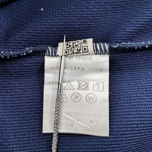 May include: Close-up of a navy blue fabric with a white tag attached. The tag has a QR code, the number "786-17D210", and washing instructions. A silver tool is holding the tag.