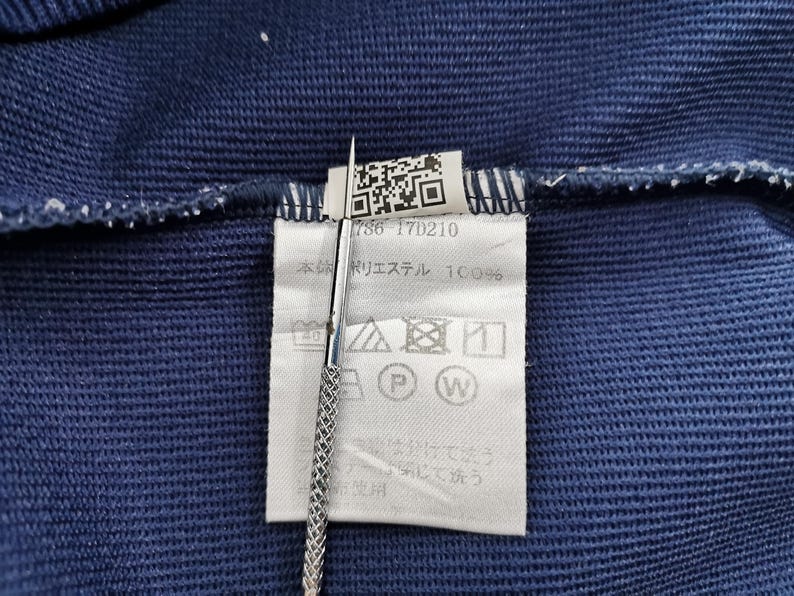 May include: Close-up of a navy blue fabric with a white tag attached. The tag has a QR code, the number "786-17D210", and washing instructions. A silver tool is holding the tag.