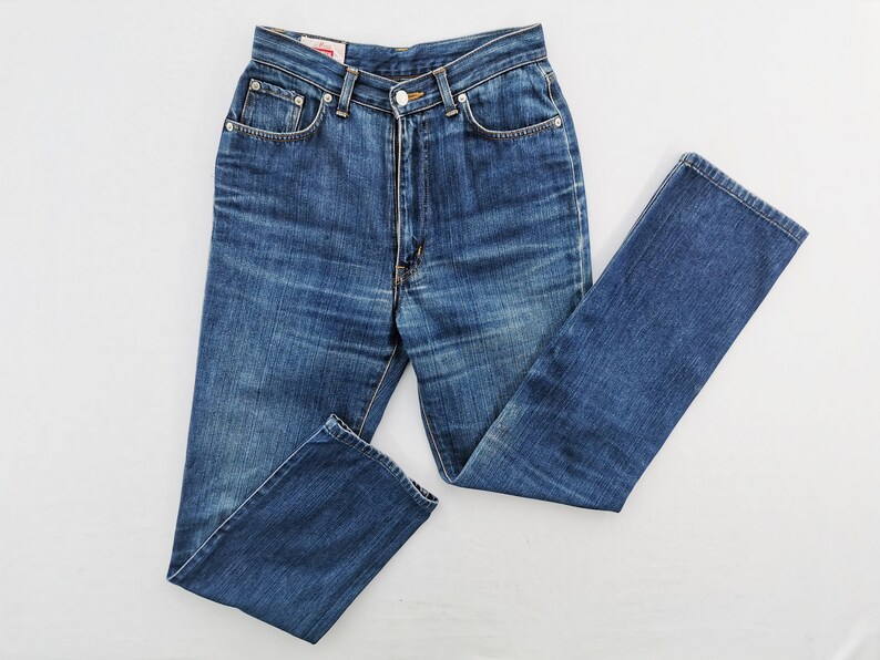 May include: Blue denim jeans with a classic five-pocket design. The jeans have a high waist and a straight leg. The fabric shows subtle fading and wear, typical of denim. The jeans are laid flat on a white surface.
