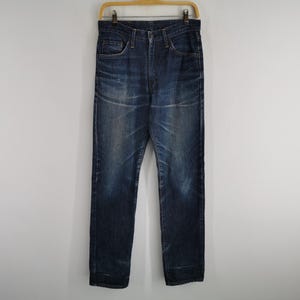 K&ouml;nnte beinhalten: Dunkelblaue Jeans, die an einem Holzb&uuml;gel h&auml;ngen. Die Jeans haben ein klassisches F&uuml;nf-Taschen-Design mit Knopf- und Rei&szlig;verschluss. Der Denim hat einen verblichenen, abgenutzten Look mit dezenten Knitterfalten und Ausbleichungen.