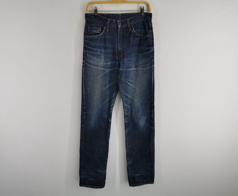 K&ouml;nnte beinhalten: Dunkelblaue Jeans, die an einem Holzb&uuml;gel h&auml;ngen. Die Jeans haben ein klassisches F&uuml;nf-Taschen-Design mit Knopf- und Rei&szlig;verschluss. Der Denim hat einen verblichenen, abgenutzten Look mit dezenten Knitterfalten und Ausbleichungen.