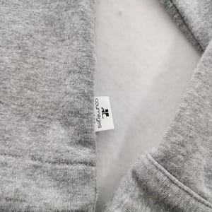 May include: Close-up of a gray sweatshirt with a white label. The label has the brand name "courr&egrave;ges" printed in black. The sweatshirt's fabric is visible, showing its texture and stitching.