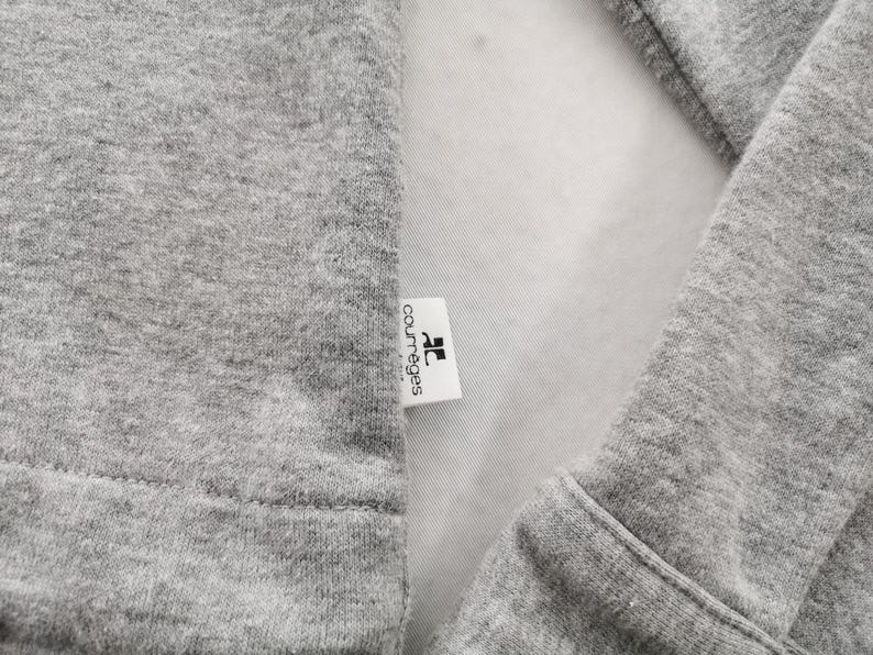 May include: Close-up of a gray sweatshirt with a white label. The label has the brand name "courr&egrave;ges" printed in black. The sweatshirt's fabric is visible, showing its texture and stitching.