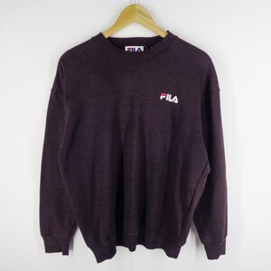 Pu&ograve; includere: Una felpa a girocollo a maniche lunghe di colore viola scuro, con il logo Fila ricamato in bianco e rosso sulla parte superiore sinistra del petto. Il capo presenta colletto, polsini e orlo a coste e include un'etichetta Fila sul collo interno.