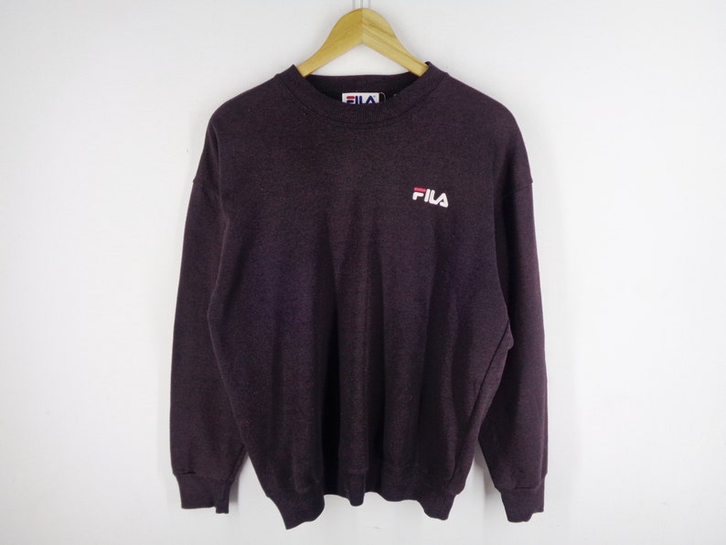 Pu&ograve; includere: Una felpa a girocollo a maniche lunghe di colore viola scuro, con il logo Fila ricamato in bianco e rosso sulla parte superiore sinistra del petto. Il capo presenta colletto, polsini e orlo a coste e include un'etichetta Fila sul collo interno.