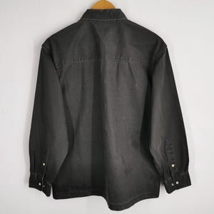 May include: A dark gray denim shirt with long sleeves, a button-down front, and a classic collar. The shirt is hanging on a wooden hanger against a white wall. The back of the shirt is visible.