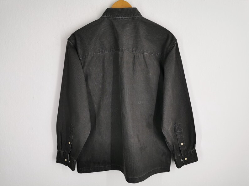 May include: A dark gray denim shirt with long sleeves, a button-down front, and a classic collar. The shirt is hanging on a wooden hanger against a white wall. The back of the shirt is visible.