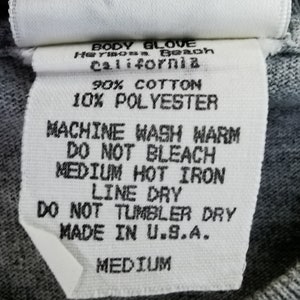 May include: A white fabric tag with black text, featuring the Body Glove logo and care instructions. The tag indicates the garment is made of 90% cotton and 10% polyester, and is a medium size. The tag also states "Made in U.S.A."