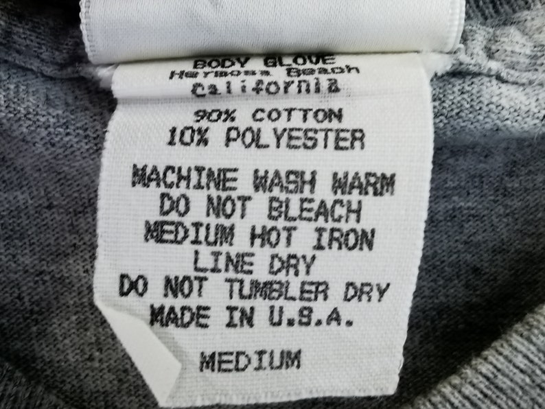 May include: A white fabric tag with black text, featuring the Body Glove logo and care instructions. The tag indicates the garment is made of 90% cotton and 10% polyester, and is a medium size. The tag also states "Made in U.S.A."