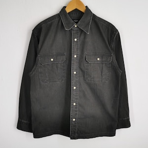 May include: A dark gray denim shirt with long sleeves, a button-down front, and two chest pockets. The shirt has a classic collar and is hanging on a wooden hanger against a white wall.