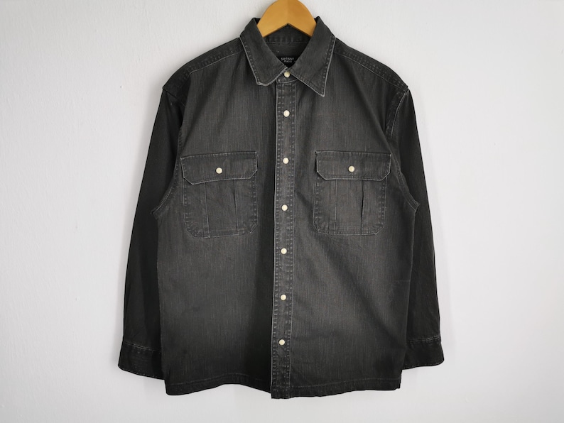 May include: A dark gray denim shirt with long sleeves, a button-down front, and two chest pockets. The shirt has a classic collar and is hanging on a wooden hanger against a white wall.