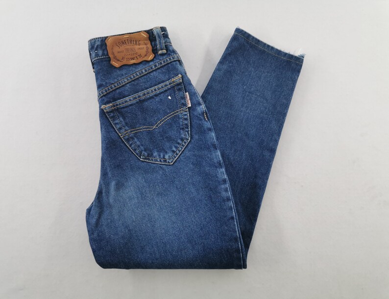 May include: Blue denim jeans with a classic five-pocket design. The jeans feature a brown leather patch on the back with the text "SOMETHING". The jeans are folded and laid flat on a white surface.