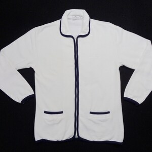 May include: White zip-up jacket with navy blue trim on the collar, cuffs, pockets, and zipper. The jacket has two front pockets and a small logo on the chest. The long-sleeved jacket is made of a soft, textured material.