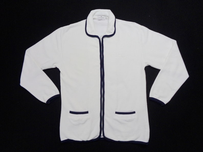 May include: White zip-up jacket with navy blue trim on the collar, cuffs, pockets, and zipper. The jacket has two front pockets and a small logo on the chest. The long-sleeved jacket is made of a soft, textured material.
