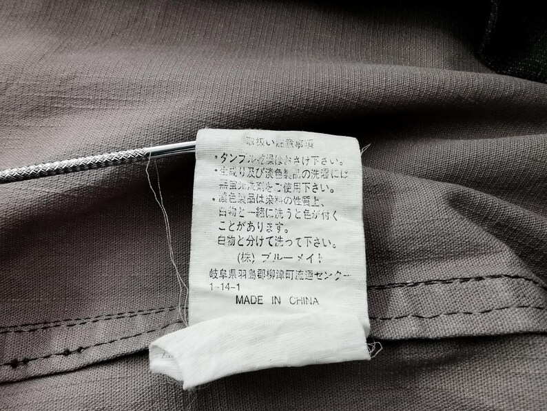 May include: Close-up of a gray fabric with a white tag attached. The tag has Japanese text and the words "MADE IN CHINA" printed in English. The fabric has a visible seam.