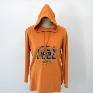 以下が含まれることがあります： オレンジ色のフード付きスウェットシャツ、長袖。前面には「ALL THAT'S JAZZ」の文字がグレーと黒でプリントされ、下に虎のグラフィックがあります。白い背景にマネキンに着せられています。