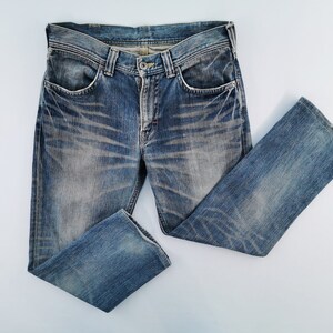 May include: Blue denim jeans with a faded wash and distressed details. The jeans have a classic five-pocket design, a button and zipper closure, and a straight leg. The denim is a medium wash with lighter areas and subtle wear marks.