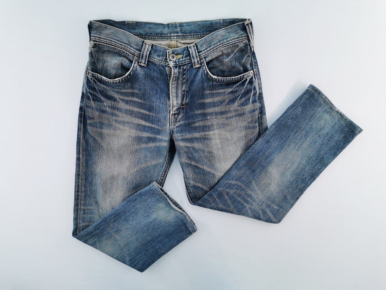 May include: Blue denim jeans with a faded wash and distressed details. The jeans have a classic five-pocket design, a button and zipper closure, and a straight leg. The denim is a medium wash with lighter areas and subtle wear marks.