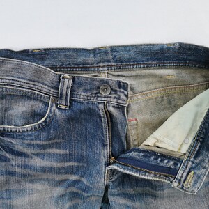 May include: Close-up of blue denim jeans with a partially unzipped fly. The jeans show signs of wear and fading, with a visible pocket and belt loops. The interior lining is a lighter color.