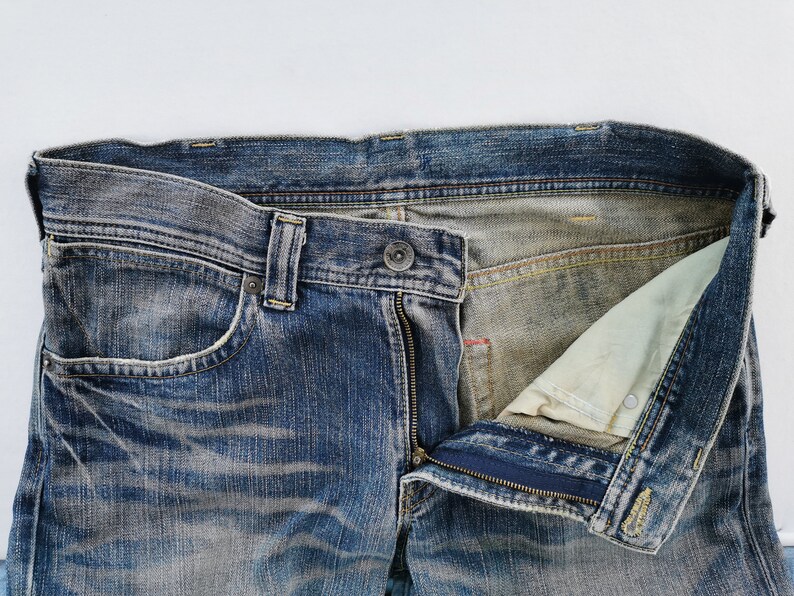 May include: Close-up of blue denim jeans with a partially unzipped fly. The jeans show signs of wear and fading, with a visible pocket and belt loops. The interior lining is a lighter color.