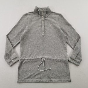 May include: A gray long-sleeve pullover shirt with a high collar and a button-down placket. The shirt has a drawstring at the waist and a small logo on the chest. The sleeves have buttoned cuffs.