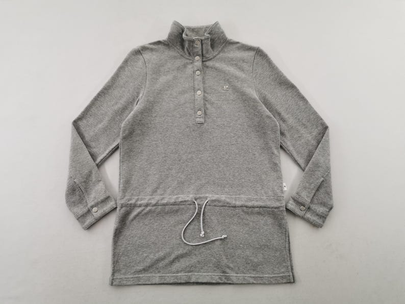 May include: A gray long-sleeve pullover shirt with a high collar and a button-down placket. The shirt has a drawstring at the waist and a small logo on the chest. The sleeves have buttoned cuffs.