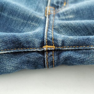 May include: Close-up of blue denim jeans, showcasing the stitching details. The image highlights the seam where the two sides of the jeans meet, with visible yellow-orange thread. The fabric has a worn, textured appearance.