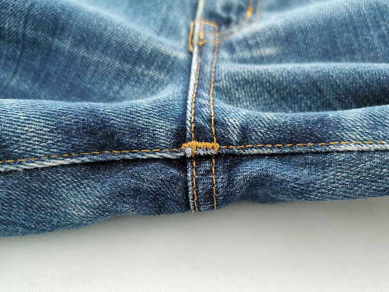 May include: Close-up of blue denim jeans, showcasing the stitching details. The image highlights the seam where the two sides of the jeans meet, with visible yellow-orange thread. The fabric has a worn, textured appearance.