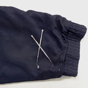 May include: Close-up of a navy blue fabric with a gathered cuff. Two silver, pointed tools are crossed over the fabric. The tools have textured handles and are used for detailed work.