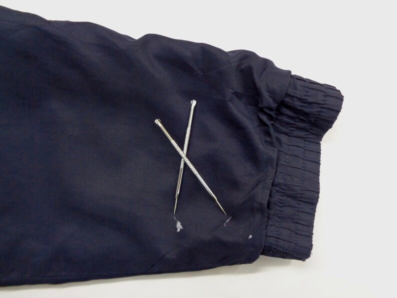 May include: Close-up of a navy blue fabric with a gathered cuff. Two silver, pointed tools are crossed over the fabric. The tools have textured handles and are used for detailed work.