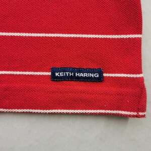Puede incluir: Primer plano de un polo rojo con rayas blancas horizontales. La camisa tiene una peque&ntilde;a etiqueta azul marino con el texto "KEITH HARING". La tela tiene una apariencia texturizada.