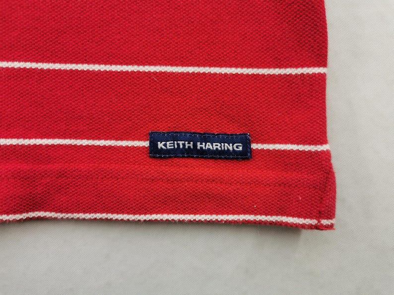 Puede incluir: Primer plano de un polo rojo con rayas blancas horizontales. La camisa tiene una peque&ntilde;a etiqueta azul marino con el texto "KEITH HARING". La tela tiene una apariencia texturizada.