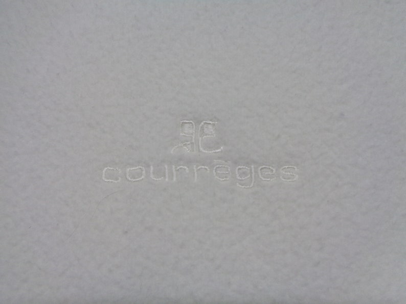 May include: A light gray textile with the embroidered word "courr&egrave;ges" in a matching light gray thread. Above the word is a stylized logo also embroidered in the same color.