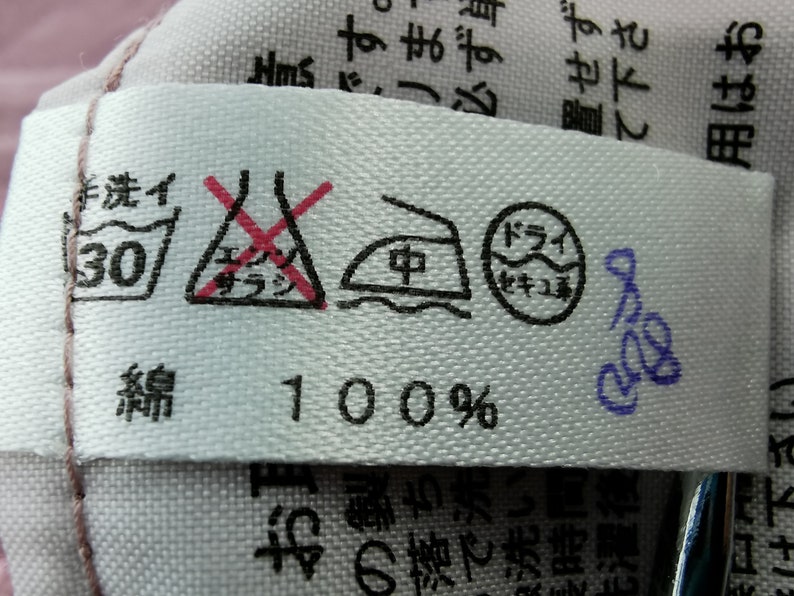 May include: A white fabric tag with care symbols and Japanese text. Symbols include a washtub with "30" inside, a crossed-out triangle, an iron, and a circle. The tag reads "100%" and has additional Japanese text.