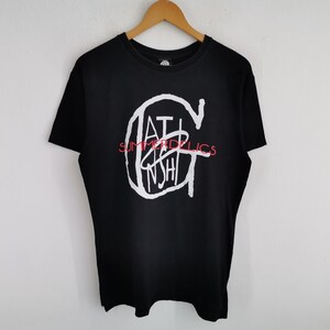 May include: A black short-sleeve t-shirt featuring a large white graphic of the letter G with the text "GATINSHI" inside it and the word "SUMMERDELICS" printed in red across the center. The shirt is displayed on a wooden clothes hanger.