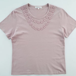 May include: A mauve-colored t-shirt with a crew neck and short sleeves. The neckline is adorned with two rows of embroidered rosettes. The tag reads "Yi for living".