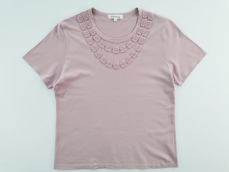May include: A mauve-colored t-shirt with a crew neck and short sleeves. The neckline is adorned with two rows of embroidered rosettes. The tag reads "Yi for living".