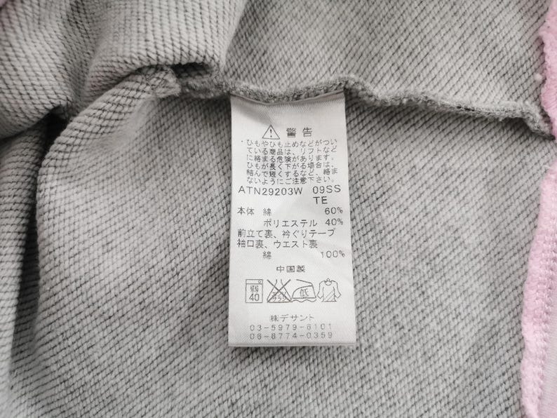 May include: Close-up of a gray sweatshirt with a pink trim. A white tag with Japanese text and care symbols is attached. The tag includes the material composition: 60% cotton, 40% polyester.