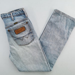 May include: Light blue Wrangler jeans with a faded wash. The jeans feature a classic five-pocket design, a brown leather patch with the Wrangler logo, and a straight leg cut. The jeans are folded neatly.