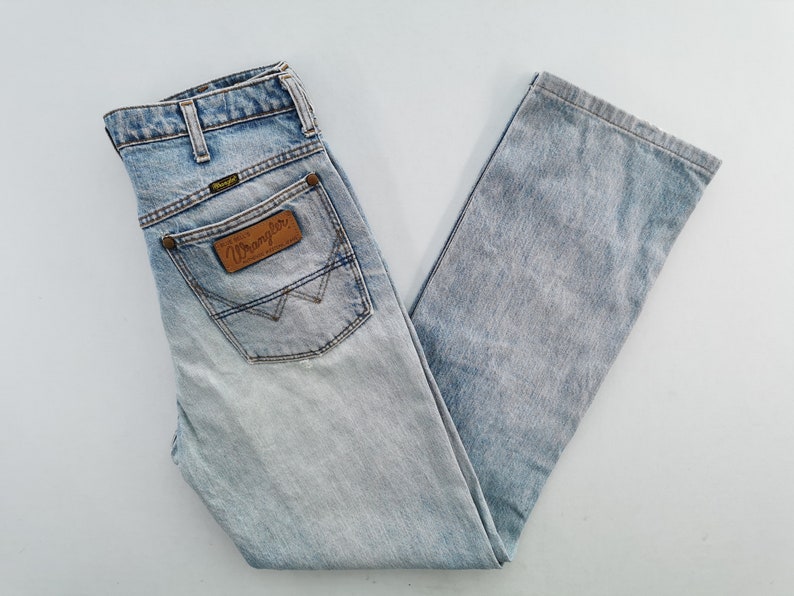 May include: Light blue Wrangler jeans with a faded wash. The jeans feature a classic five-pocket design, a brown leather patch with the Wrangler logo, and a straight leg cut. The jeans are folded neatly.
