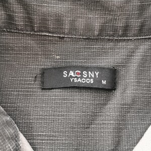 May include: Close-up of a gray collared shirt with a textured pattern. A black label with the text "SACSNY Y'SACCS M" is sewn on the shirt. The shirt is made of a woven fabric.